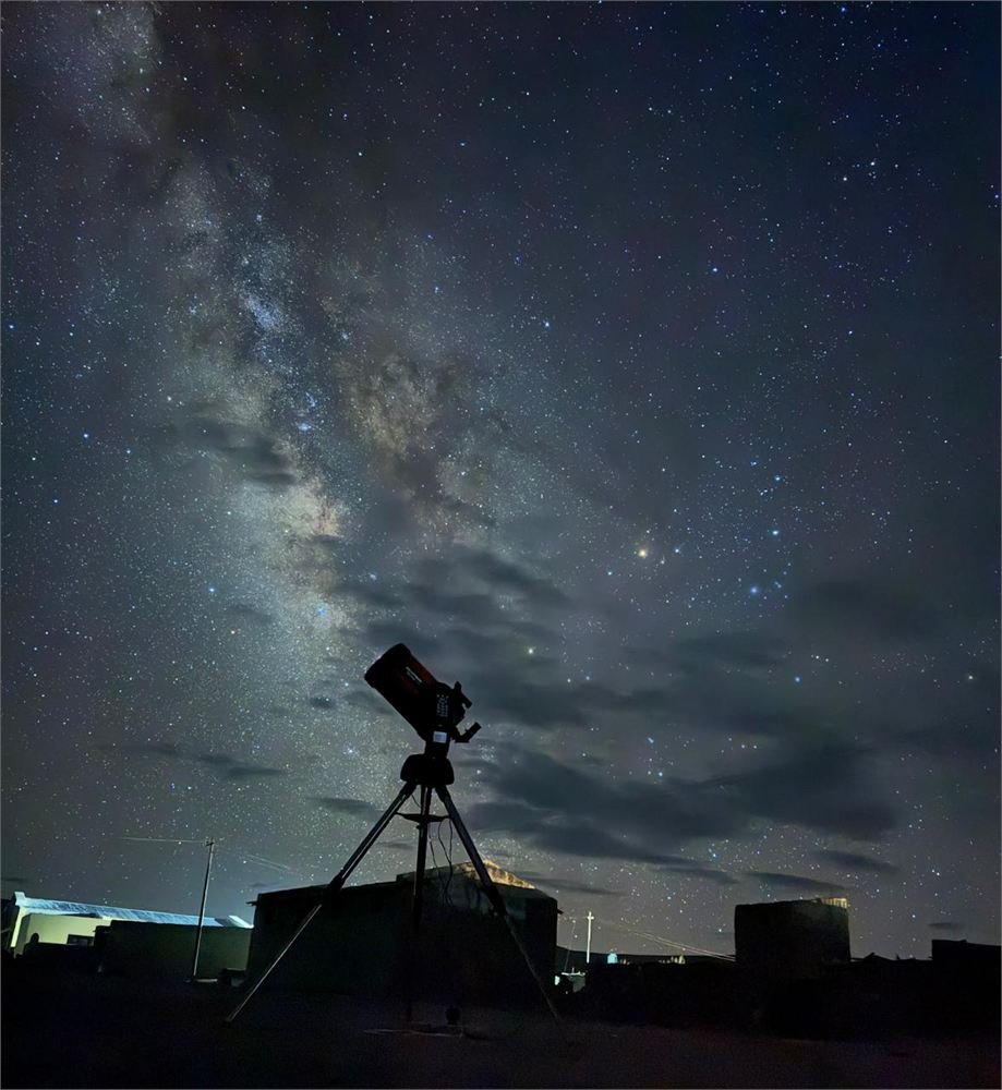 A telescope on a tripod silhouetted against the Milky Way rising over Ladakhi rooftops